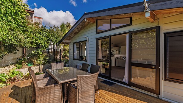 A patio with a glass table and wicker chairs, a wooden deck, and a house with sliding glass doors. - Doubtless Bay Vista - Cable Bay Retreat (Cable Bay)