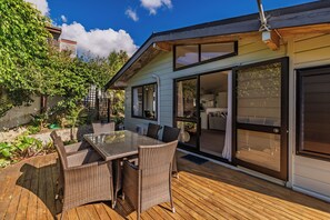 A patio with a glass table and wicker chairs, a wooden deck, and a house with sliding glass doors. - Doubtless Bay Vista - Cable Bay Retreat (Cable Bay)