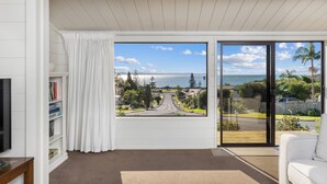 A living room with a large window offering a view of a road and greenery, a white sofa, and a bookshelf. - Doubtless Bay Vista - Cable Bay Retreat (Cable Bay)