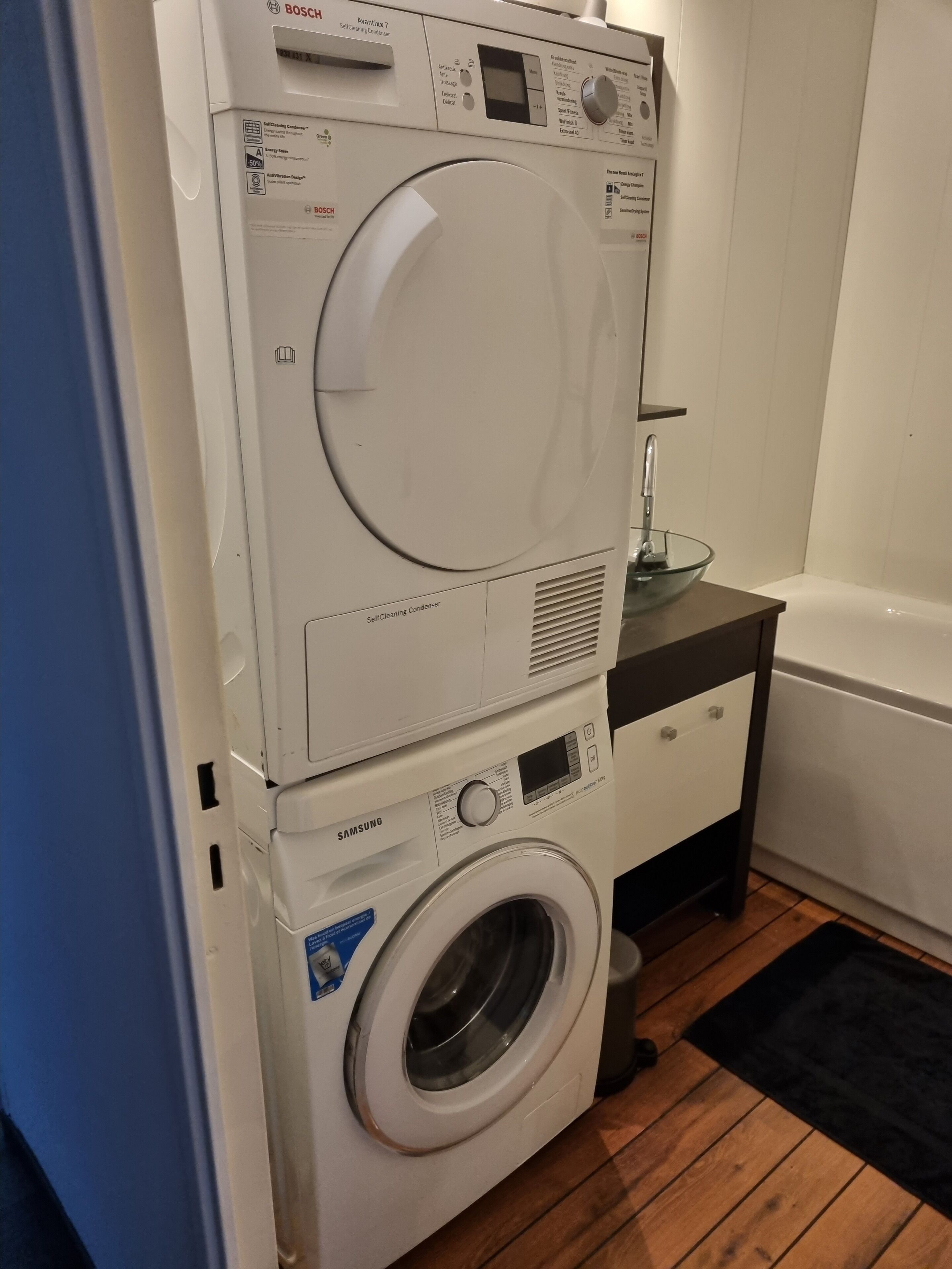 Un appareil combiné lave-linge et sèche-linge, installé en hauteur, dans une salle de bain avec un plancher en bois et un lavabo en verre.