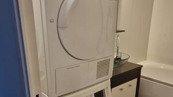 Un appareil combiné lave-linge et sèche-linge, installé en hauteur, dans une salle de bain avec un plancher en bois et un lavabo en verre.