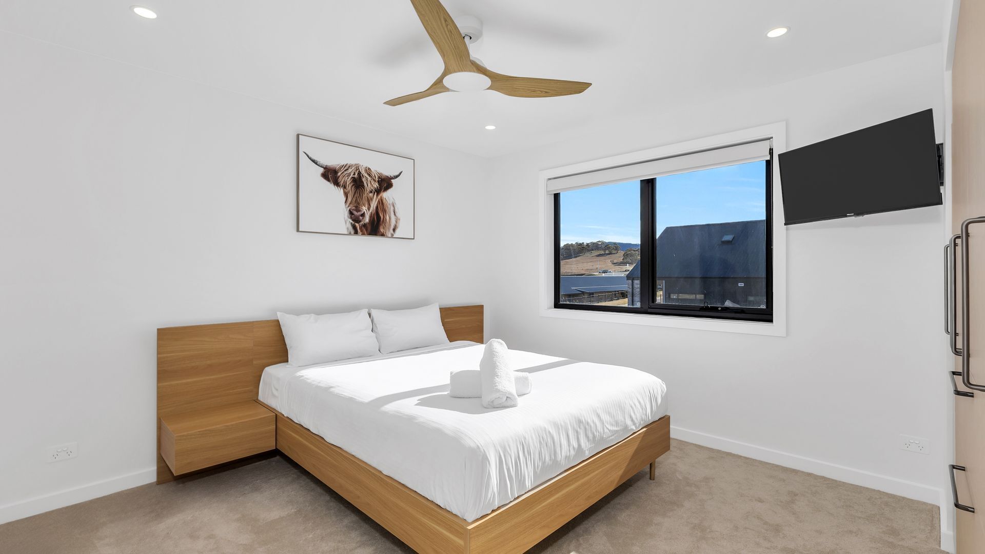 A bedroom with a bed, a ceiling fan, a window with a view, and a mounted TV.