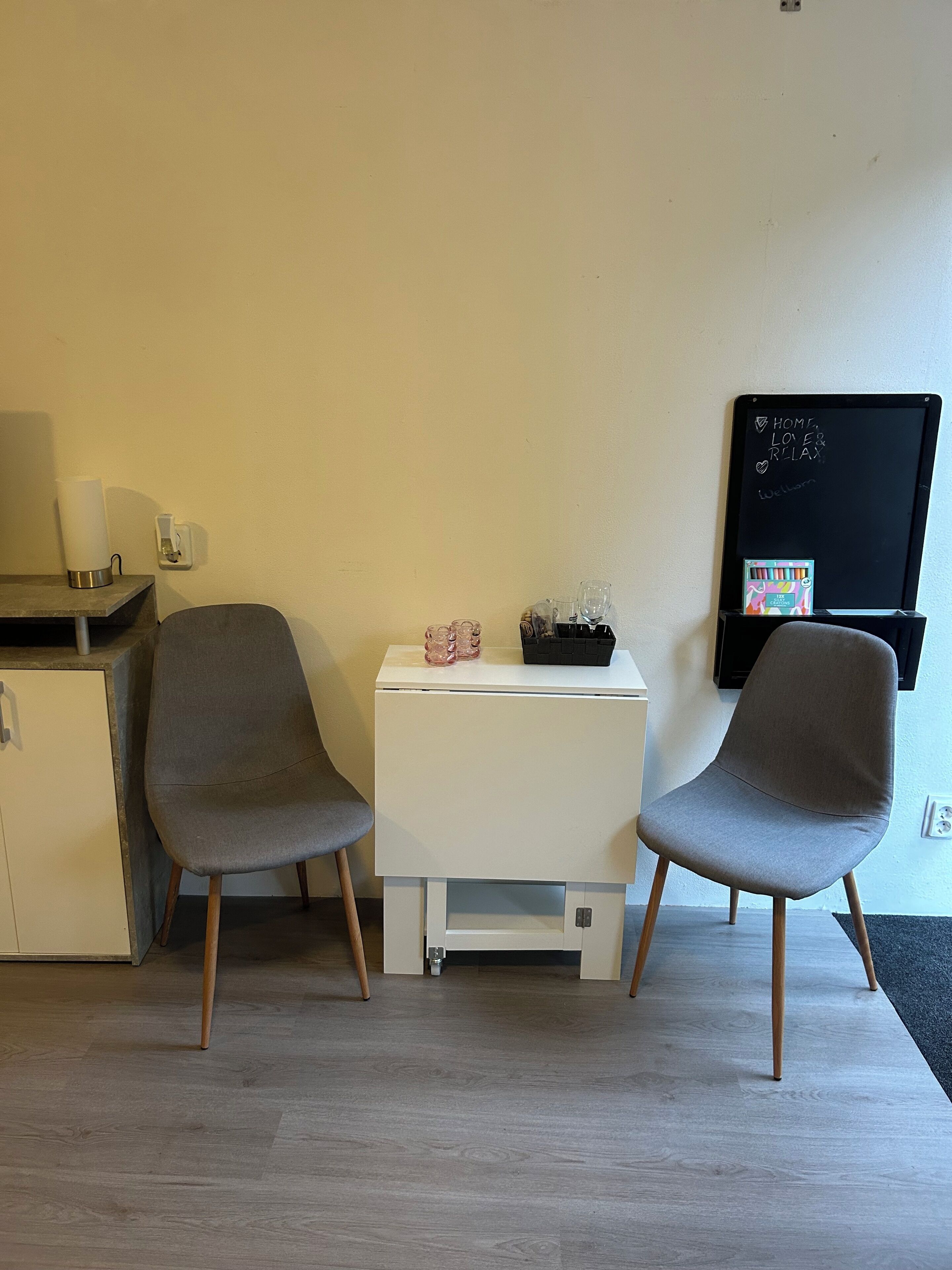 A room with two grey chairs, a small white table, a blackboard with motivational writing, and a desk with a lamp and a water bottle.