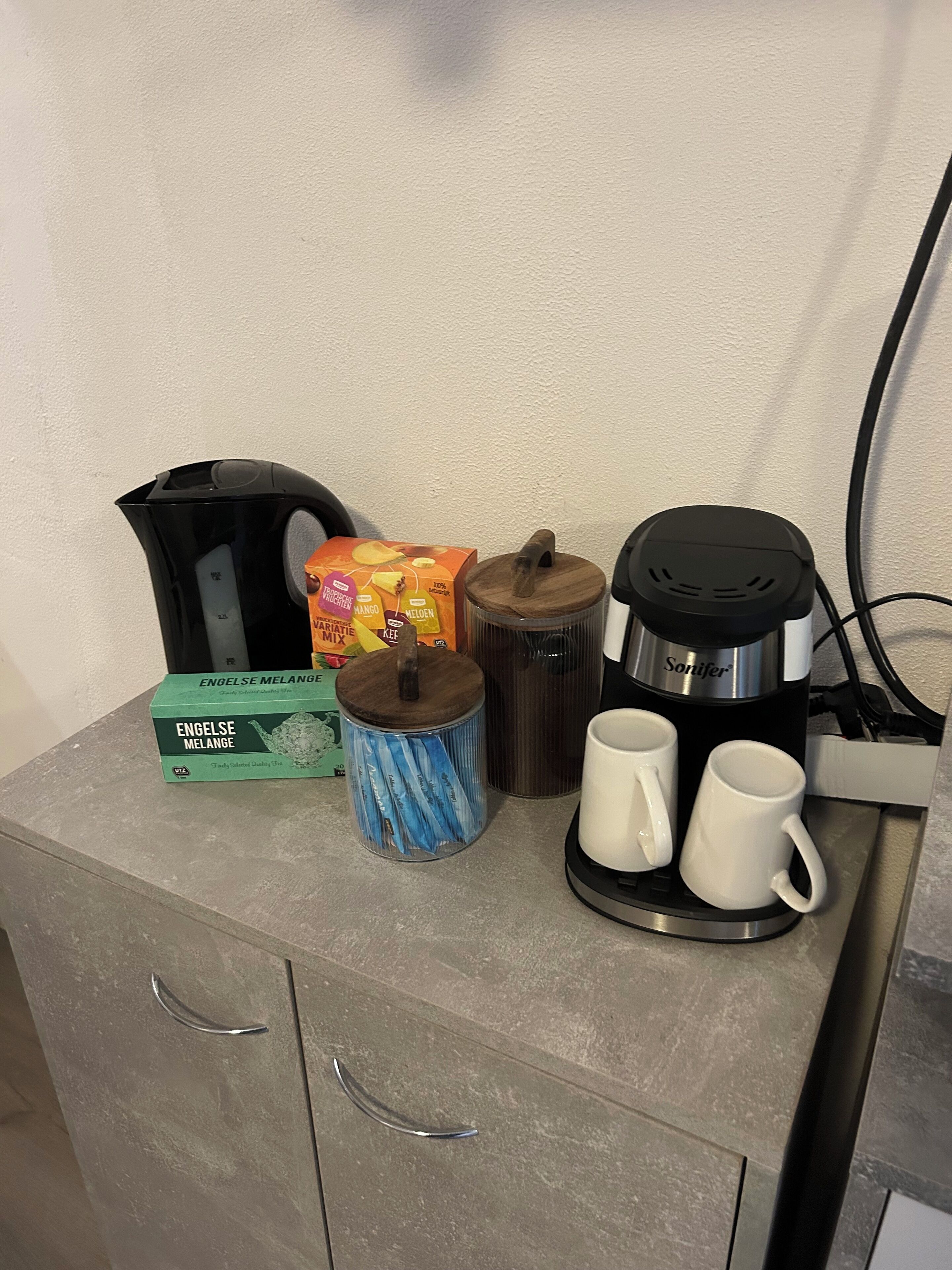 Ett litet kök med en kaffebryggare, en ask te, en mugg och en behållare.