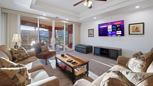 Un salon moderne doté d’un ventilateur de plafond, d’un grand téléviseur à écran plat diffusant des services de streaming et offrant une vue à travers une porte-fenêtre coulissante.