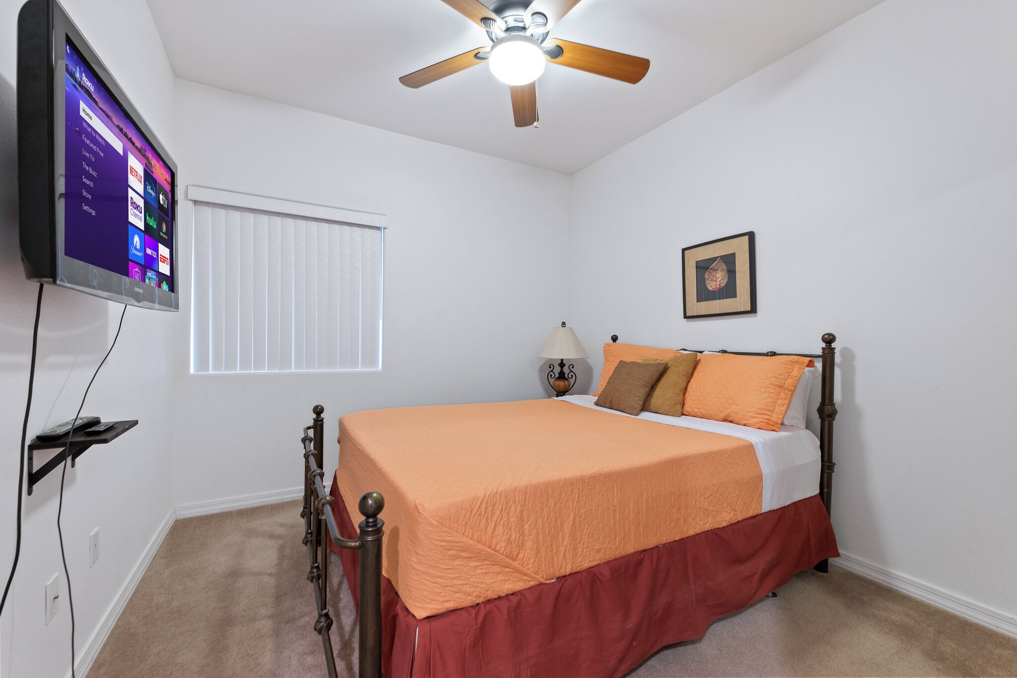 Um quarto com cama, ventilador de teto, TV fixada na parede, janela com cortinas e mesa de cabeceira com abajur.