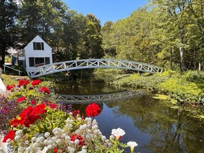 Ett vitt hus med en liten veranda, en träbro över en damm och färgglada blommor i förgrunden.