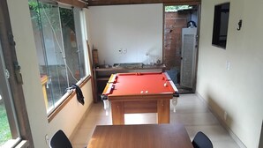 Una mesa de billar en una habitación con una mesa de madera, sillas negras, ventanas amplias y una pared de ladrillo.