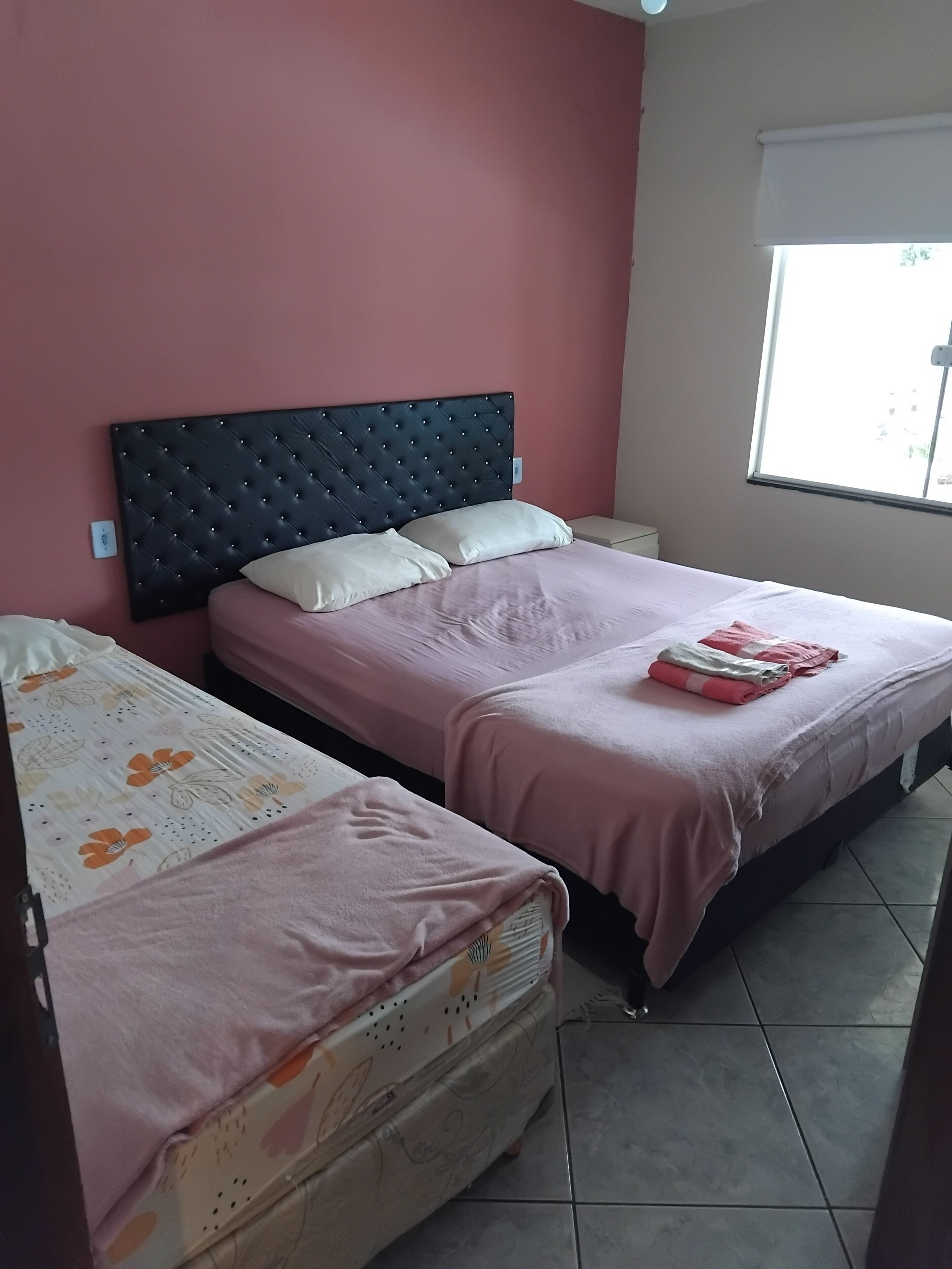A room with two beds, each with a pink bedspread and a headboard. There is a window with a view of the outside.