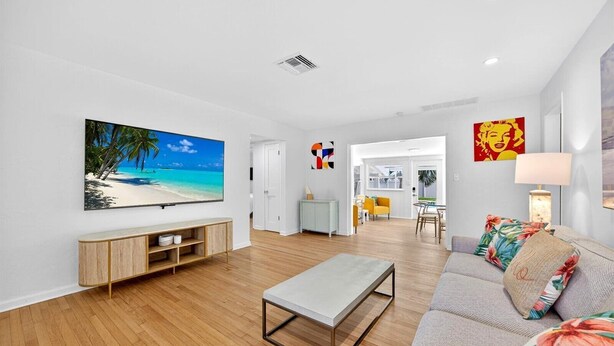 A modern living room with a flat-screen TV, wooden flooring, a grey sofa, and a dining area in the background.