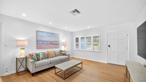 A spacious living room with a grey sofa, two side tables with lamps, a coffee table, a large painting of a beach, and a window with blinds.