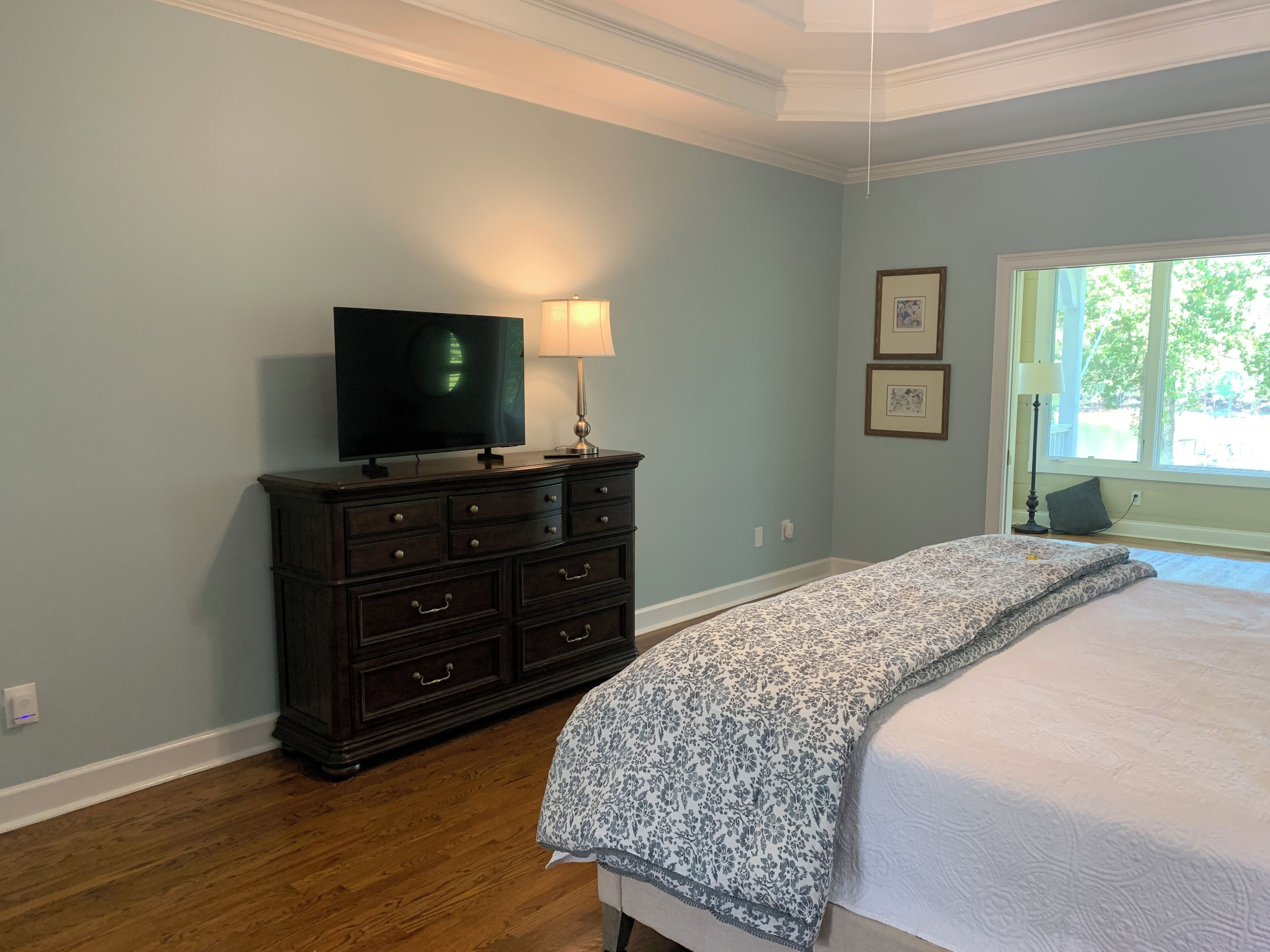A bedroom with a bed, a dresser, a television, and a window with a view of greenery.