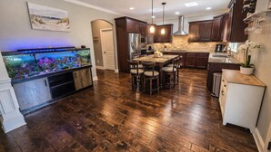 A kitchen with wooden flooring, dark cabinets, a built-in fish tank, pendant lights, and a dining area with chairs.