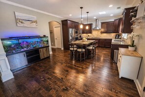 Una cocina con suelo de madera, armarios oscuros, un acuario integrado, luces colgantes y una zona de comedor con sillas.
