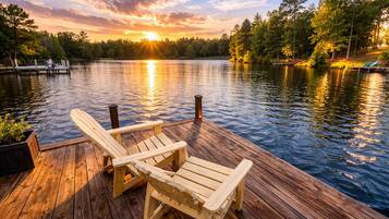 A wooden dock with two Adirondack chairs facing a calm lake at sunset.