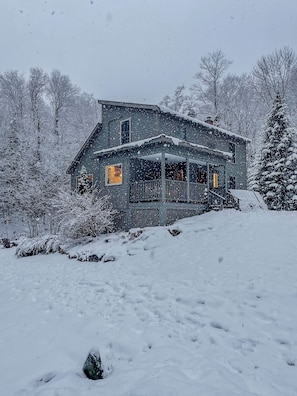 Una casa de madera de dos pisos con un porche, rodeada de árboles cubiertos de nieve.