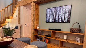 Una sala de estar acogedora con una escalera de madera, un cuadro grande de un bosque y una estantería con diversos objetos.