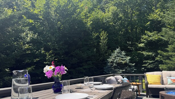 Un área de comedor al aire libre con terraza de madera, una mesa dispuesta con platos, vasos y un jarrón de flores.