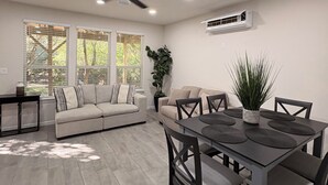 A modern living room with a sofa, dining table, and chairs. There are windows with blinds, a ceiling fan, and an air conditioning unit on the wall. - Cozy Casita in San Antonio Close to sea world- downtown-fiesta Texas-Alamo ranch (San Antonio)