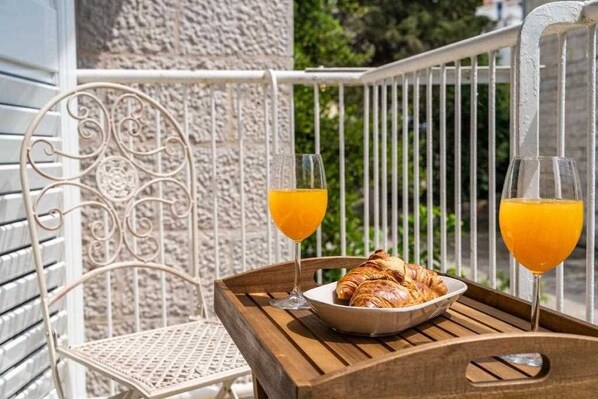 A balcony with a wicker chair, a wooden table, a bowl of croissants, and two glasses of orange juice.