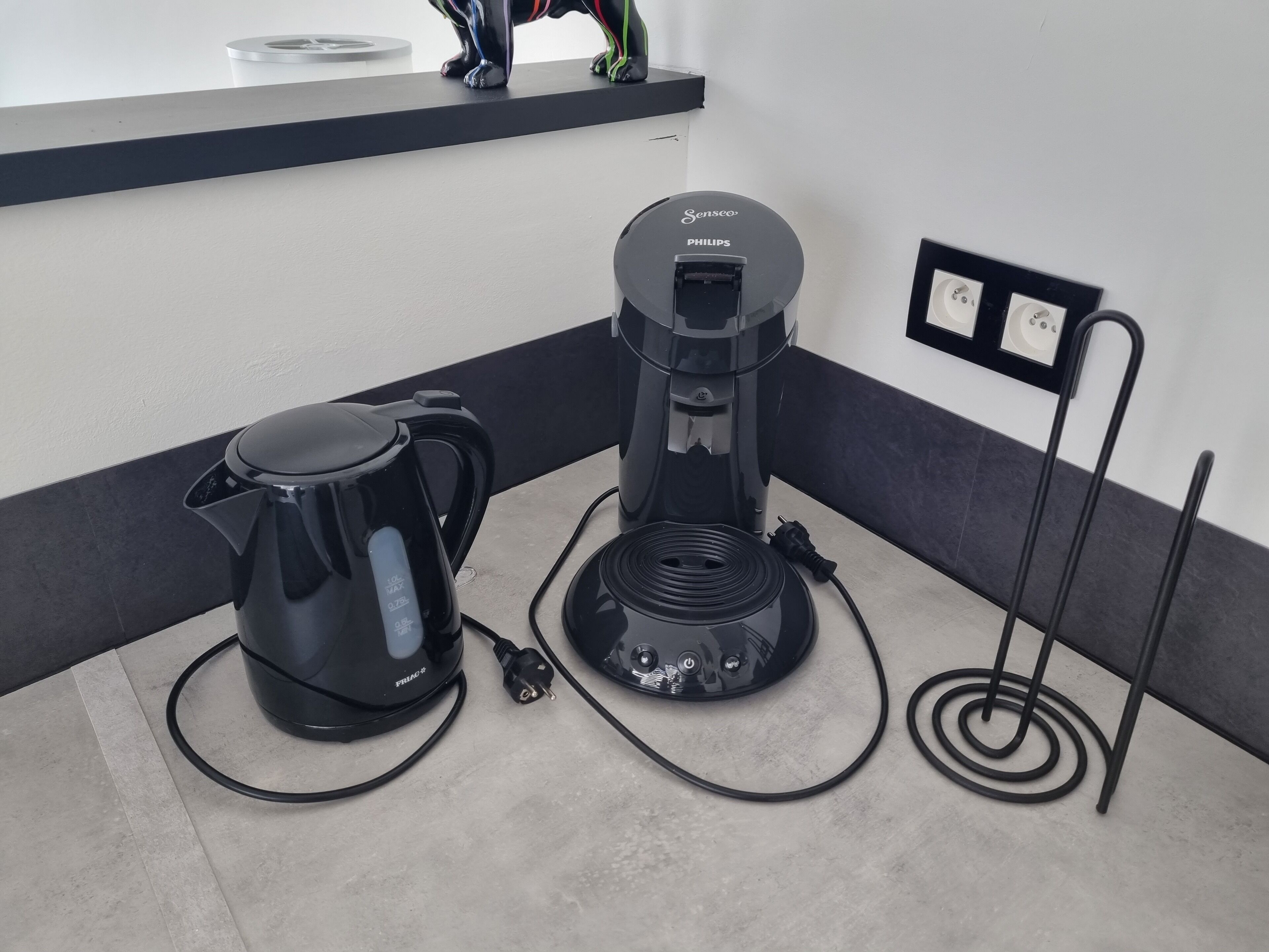 Une machine à café et une bouilloire sont posées sur un plan de travail, à proximité d’une prise de courant et d’un interrupteur fixé au mur.