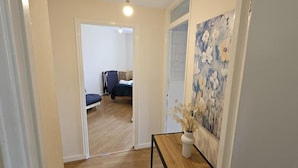 Corredor que dá para um quarto com uma cama, mesa de cabeceira e um vaso com flores secas.