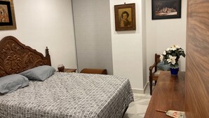 A bedroom with a wooden headboard, a patterned bedspread, a nightstand with a vase, a wooden chair, and a table with a vase of flowers.