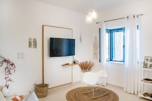 A modern living room with a flat-screen TV, a white chair, a wooden rocking chair, a woven basket, and a window with blue curtains.