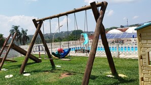 Un parque infantil con columpios y un tobogán, una piscina con cerramiento blanco, y paneles solares al fondo.
