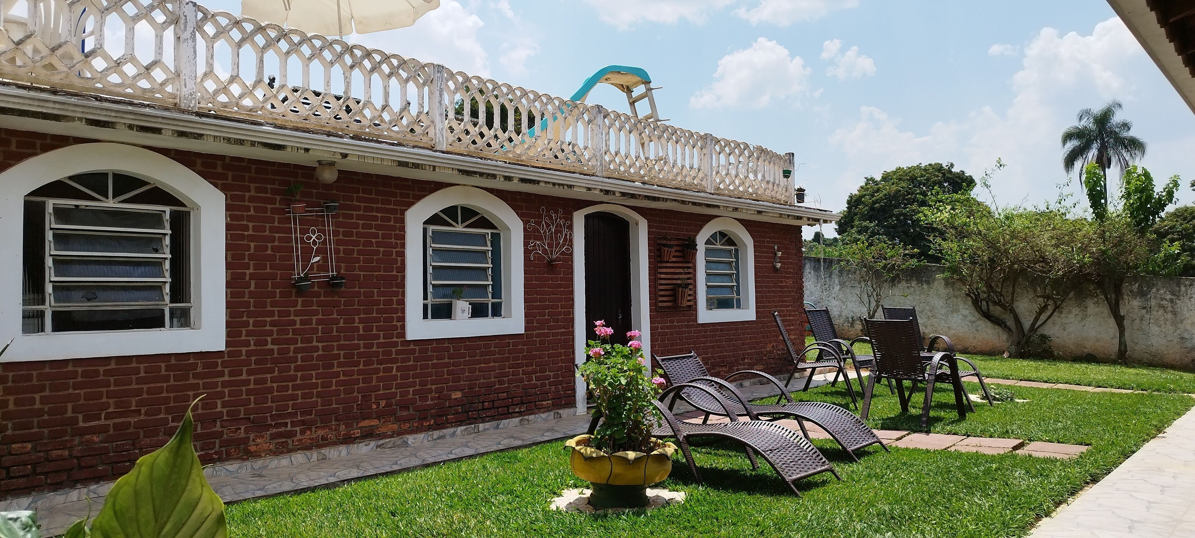 Uma casa de tijolos com uma varanda, um jardim com cadeiras e uma palmeira.