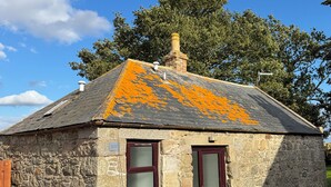 A stone cottage with a sloped roof, two windows with dark frames, and a chimney.