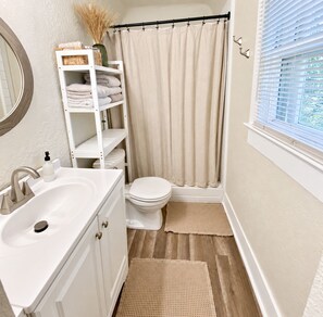 Une salle de bain avec un lavabo blanc, des toilettes et un rideau de douche. On y trouve une étagère avec des serviettes et un vase contenant des plantes séchées.
