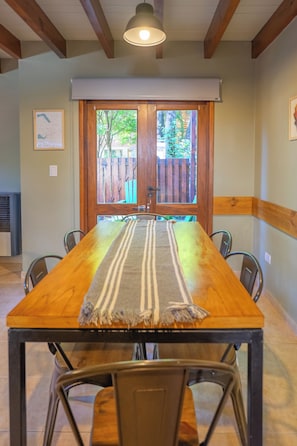 A dining area with a long wooden table, six chairs, a striped table runner, and a window with a view of a wooden fence and greenery.