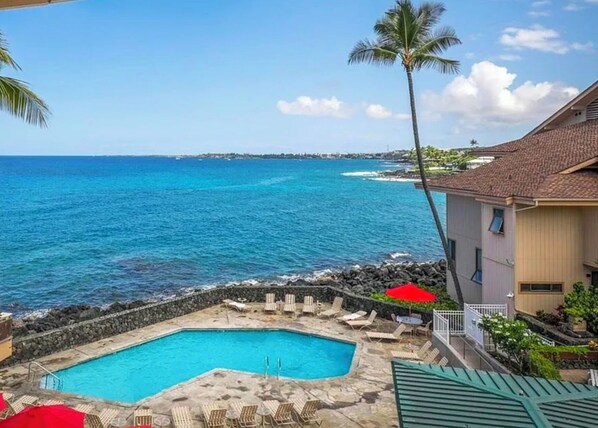 Un espace piscine doté de chaises longues et de parasols, avec vue sur une plage et l’océan.