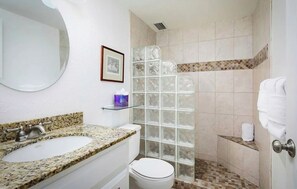 Une salle de bain avec un lavabo surmonté d’un plan de travail en granit, des toilettes blanches, une douche en verre et un tableau encadré au mur.