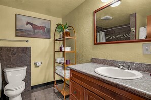 Une salle de bain comprenant des toilettes, un lavabo, un meuble-lavabo en bois, un porte-serviettes, une étagère avec des articles de toilette, un miroir et un tableau représentant un cheval.