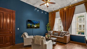 Ein Wohnzimmer mit blauer Wand, Holzboden, Deckenventilator, einem großen Fenster mit Vorhängen, einem Sofa, einem Couchtisch und einem Fernseher an der Wand.