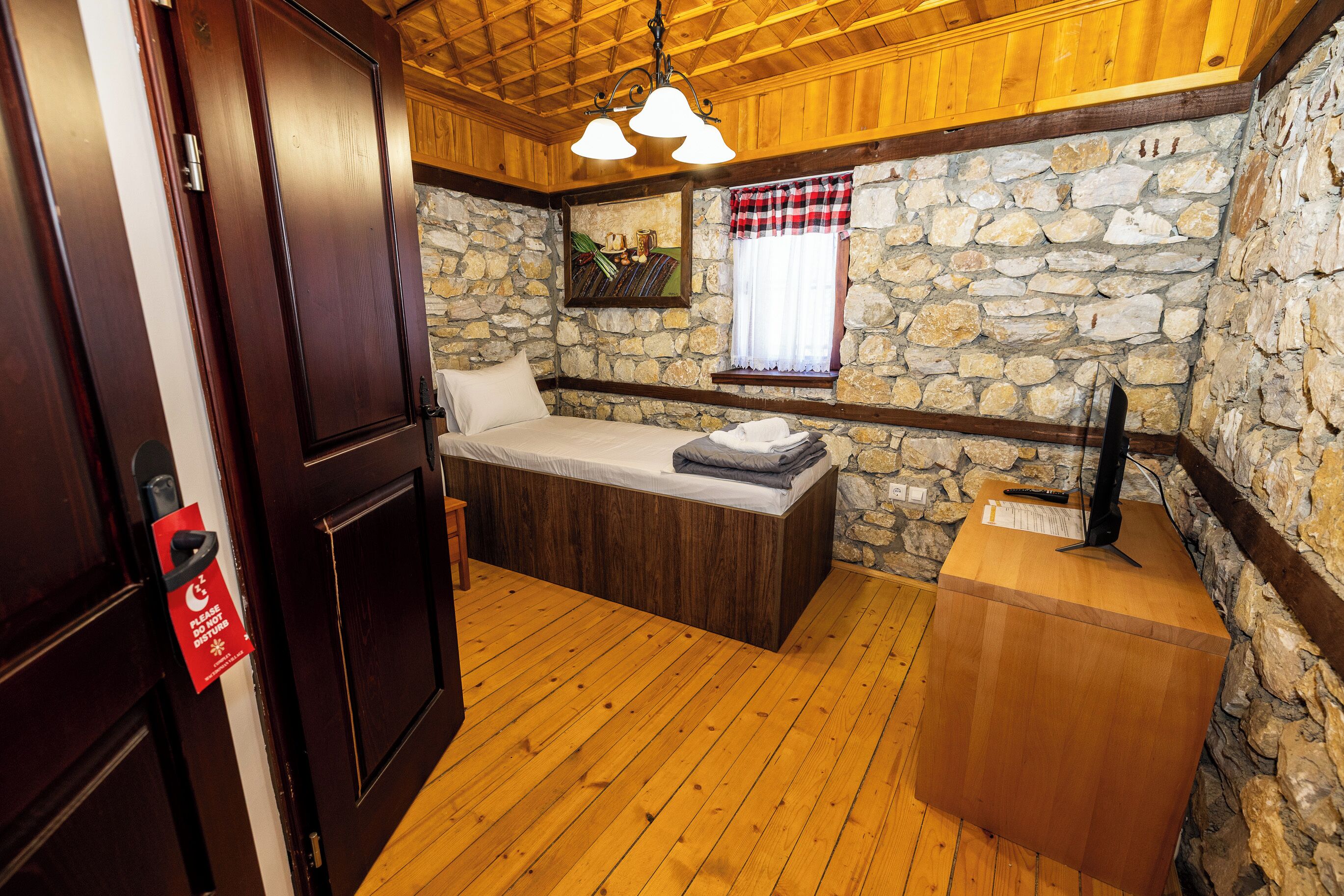 Une pièce avec un lit, un mur en pierre, un plafond en bois et une petite fenêtre ornée d’un rideau à carreaux.