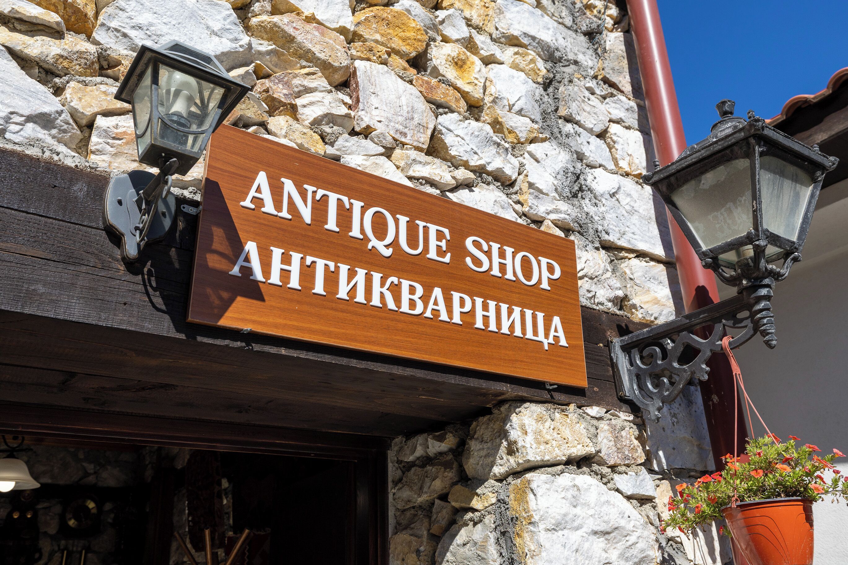 A wooden sign for an antique shop with a stone wall background and two hanging lanterns.