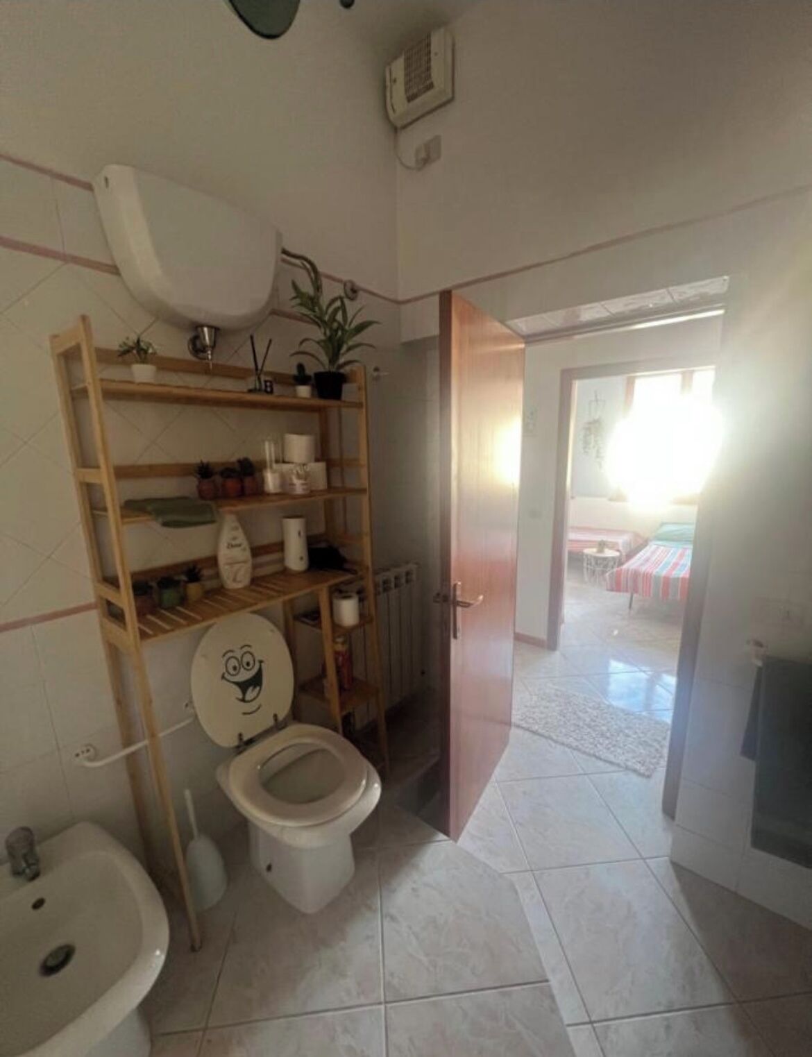 Un baño con inodoro, lavabo, repisas con artículos de aseo, un radiador y una puerta que da a otra habitación.