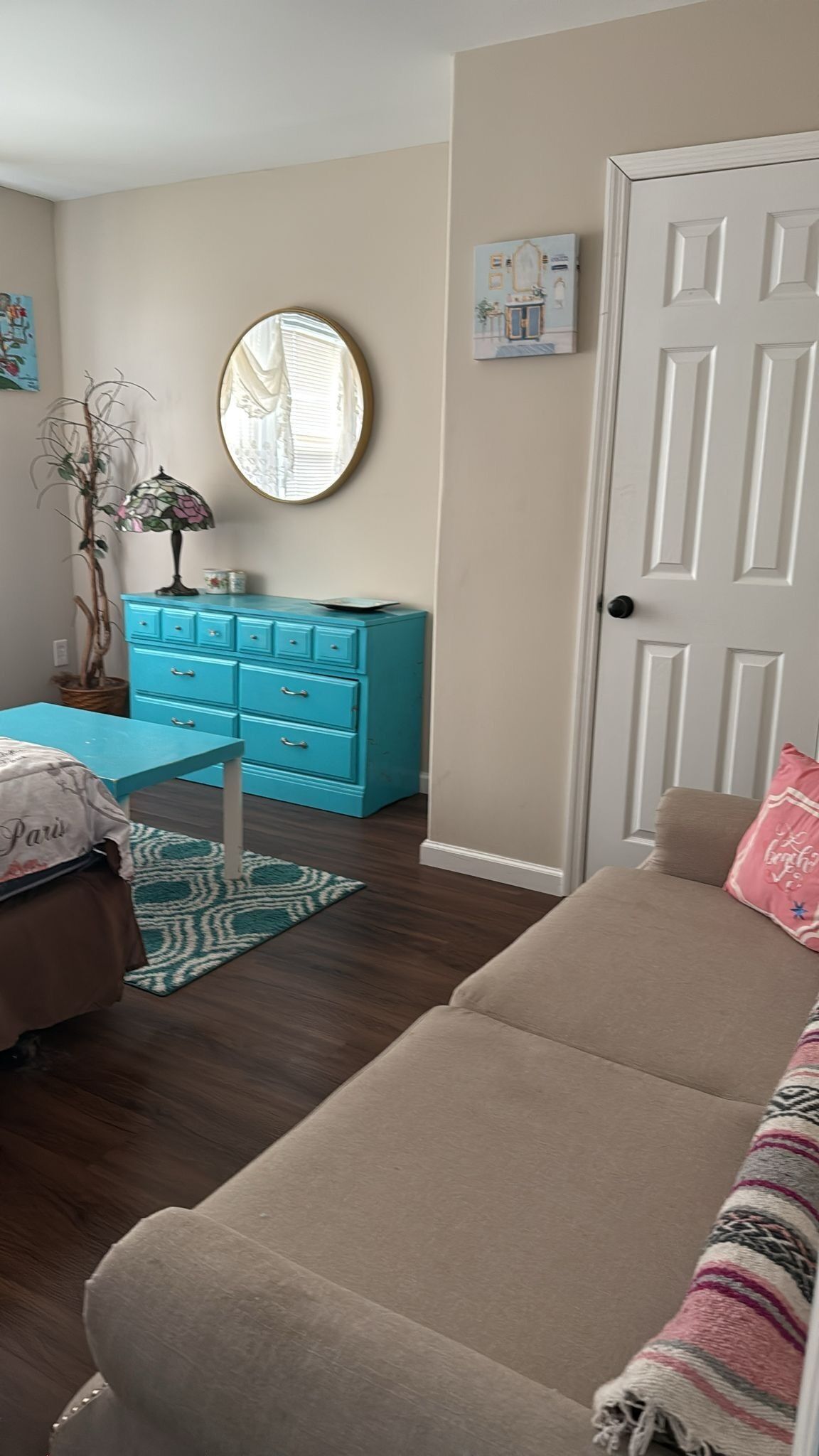 A room with a beige sofa, a blue dresser, a round mirror, and a door with a small picture.