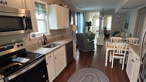 A modern kitchen with stainless steel appliances, a dining area with white chairs, and a living room with a sofa and a coffee table.