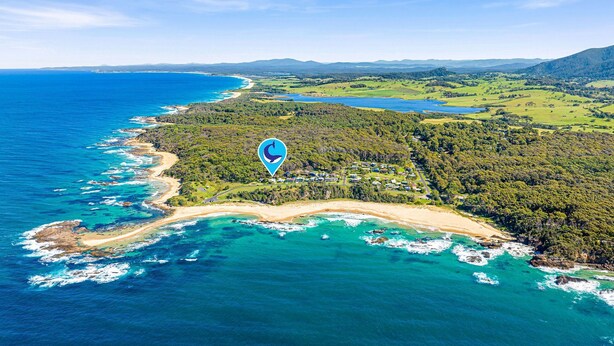 Vista aérea de una zona costera con playa, colinas boscosas y un mar azul y despejado.
