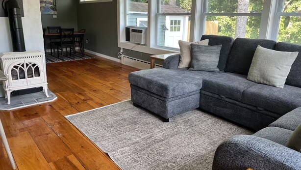 Un salon chaleureux avec un canapé d’angle, un poêle à bois blanc et de grandes fenêtres donnant sur l’extérieur.