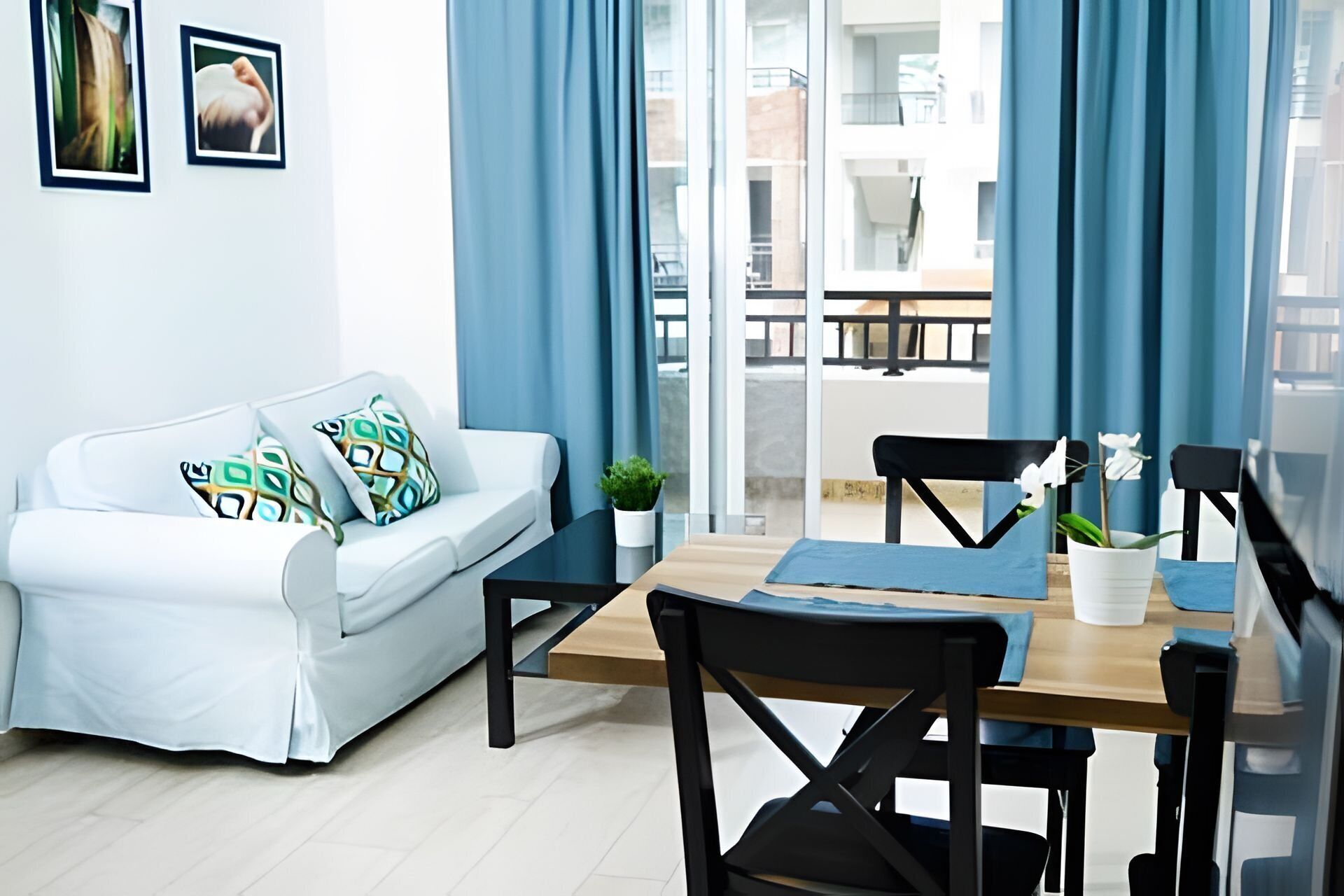 Ein modernes Wohnzimmer mit einem weißen Sofa, zwei gemusterten Kissen, einem kleinen Tisch mit einer Pflanze und einem Esstisch mit Stühlen.