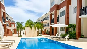 A swimming pool with lounge chairs and umbrellas, surrounded by multi-story buildings with balconies. - Condo with Rooftop steps from the beach! (Punta Cana)