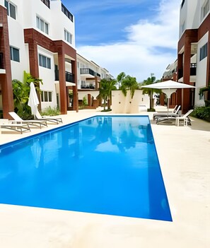 Ein Schwimmbecken mit Liegestühlen und Sonnenschirmen, umgeben von mehrstöckigen Gebäuden mit Balkonen.
