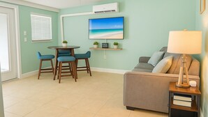 Ein Wohnzimmer mit grauem Sofa, einem runden Tisch mit blauen Stühlen, einem Fernseher an der Wand und einer Lampe auf einem Beistelltisch.