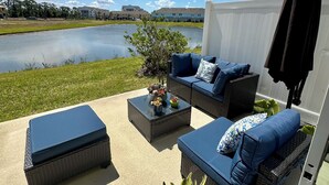 Un patio al aire libre con mobiliario de mimbre, una pequeña mesa con una planta y vistas a un lago y edificios al fondo.