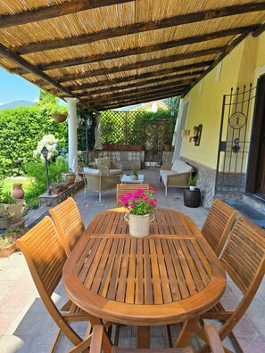 Un patio couvert comprenant une table ronde en bois et quatre chaises, entouré de plantes en pot et d’un canapé en osier.